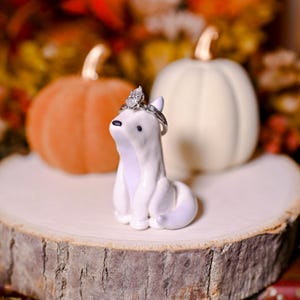 May include: A white ceramic fox ornament with a silver ring on its head, displayed on a wooden slice. Two pumpkins, one orange and one cream, are in the background. The ring features a pear-cut diamond.