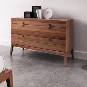 May include: A three-drawer dresser made of wood with a light brown finish. The dresser has a simple design with small, rectangular handles on each drawer. The dresser is sitting on a light gray floor.