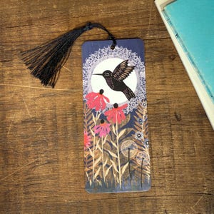 May include: A black hummingbird with a long beak flies in front of a full moon with a white lace pattern. The hummingbird is surrounded by pink and purple flowers and green leaves on a dark blue background. The bookmark has a black tassel.