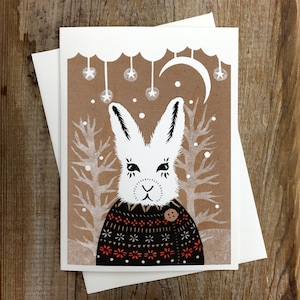 Peut inclure: Un lapin blanc portant un pull à motifs noirs et rouges avec un bouton sur le devant. Le lapin se tient dans une forêt enneigée avec des arbres et une lune croissante en arrière-plan.