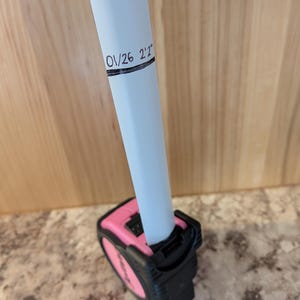 May include: A pink and black tape measure with a white pole inserted. The pole has the text "01/26 2'2"" written on it. The tape measure is on a light-colored surface.