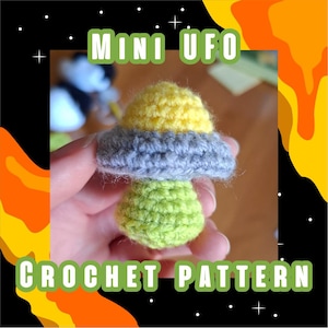 May include: A handmade crochet UFO in yellow, gray, and green. The text "MINI UFO" and "CROCHET PATTERN" are displayed in green. The UFO is held in a hand against a black background with orange and yellow accents.