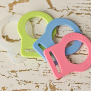 May include: Three silicone bottle rings in different colors: light green, light blue, and pink. Each ring has a small handle and a circular opening for a bottle.