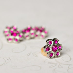 May include: A silver-toned ring with a flower design, featuring bright pink gemstones. The ring is in focus, with a matching pair of earrings slightly blurred in the background. The jewelry is set against a white, patterned surface.