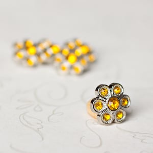 May include: A silver flower-shaped button with yellow rhinestones. The button is on a white surface with a floral pattern.