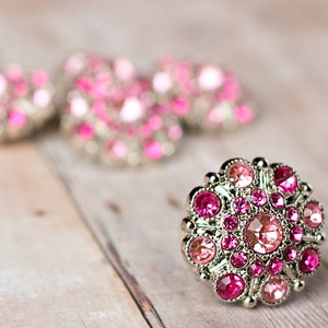 May include: A close-up of four pink and silver jeweled buttons. The buttons are round and have a raised design. The buttons are on a light brown wooden surface.