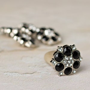 May include: A silver ring with a cluster of black and clear rhinestones. The ring has a round shape and is set on a white background.