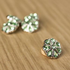 May include: Close-up of three flower-shaped earrings with green gemstones and silver-colored metal. The earrings are set against a light brown wooden surface. The earrings are in focus, with the background slightly blurred.