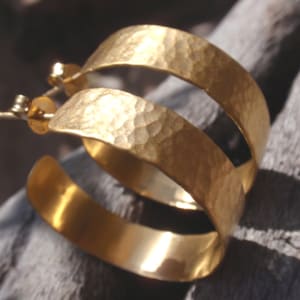 Könnte beinhalten: Goldene gehämmerte Creolen mit einem Doppelband-Design. Die Ohrringe sind aus dickem, strukturiertem Metall gefertigt und haben eine polierte Oberfläche.