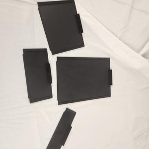 Puede incluir: Cuatro carpetas de archivos negras con una pestaña en la esquina superior derecha. Las carpetas están sobre una superficie blanca.