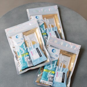 Op de afbeelding: Drie doorzichtige plastic zakjes met gouden accenten, elk met een assortiment kleine, individueel verpakte artikelen. De zakjes zijn gelabeld met "Travel Kit" en dragen de merknaam "Liquid I.V."