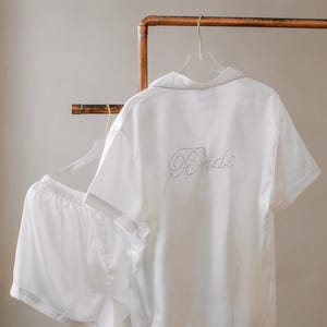 May include: White satin pyjama set with short sleeves and shorts. The top has "Bride" written on the back in rhinestones.