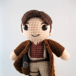 May include: A crocheted plush toy of a person wearing a brown coat, brown pants, and brown boots. The toy has brown hair and a smiling face with brown eyes.