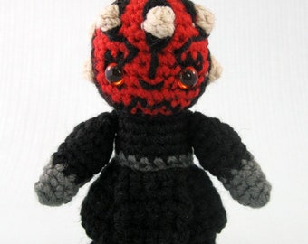 May include: A handmade crochet doll of a character with a red face, black markings, and small horns. The doll has large orange eyes, a black outfit, and gray accents. The doll is a unique, handcrafted item.