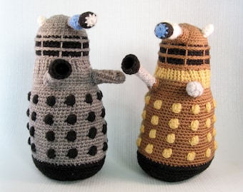 May include: Two crocheted plush toys shaped like Daleks from the Doctor Who television series. The Dalek on the left is gray with black dots and the Dalek on the right is brown with yellow dots.