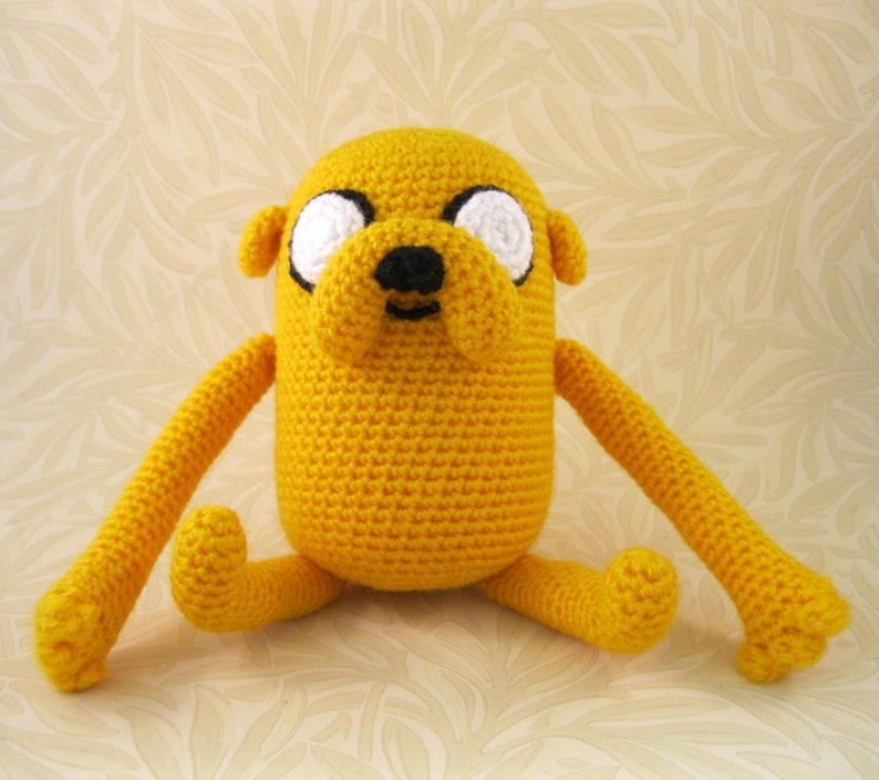 May include: A yellow crocheted stuffed dog with white eyes and a black nose. The dog is sitting with its legs outstretched.