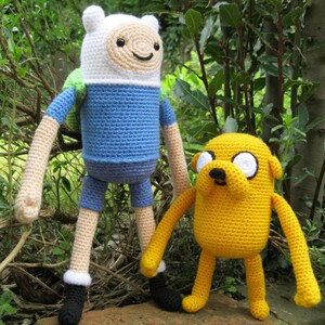 K&ouml;nnte beinhalten: Geh&auml;kelte Pl&uuml;schtiere von Finn der Mensch und Jake der Hund aus der Zeichentrickserie Adventure Time. Finn tr&auml;gt ein blaues Hemd und eine wei&szlig;e M&uuml;tze, w&auml;hrend Jake gelb mit einer schwarzen Nase ist.