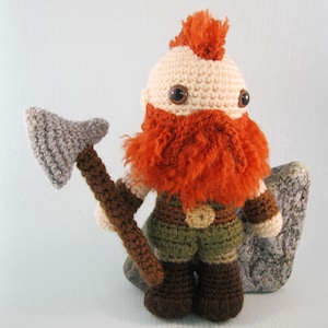 May include: A crocheted plush toy of a red-haired dwarf with a brown beard, wearing a green and brown tunic and brown boots. The dwarf is holding a grey and brown axe.