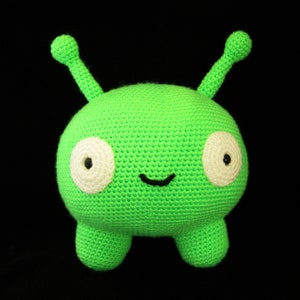 May include: A green crocheted plush toy shaped like an alien with large white eyes and a small black mouth. The toy has two small arms and two small legs.