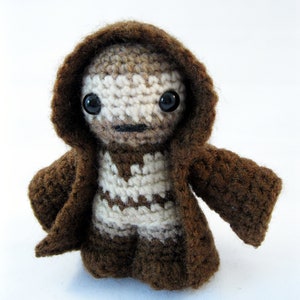 May include: A crocheted brown hooded robe with a white crocheted figure inside. The figure has black eyes and a brown face.