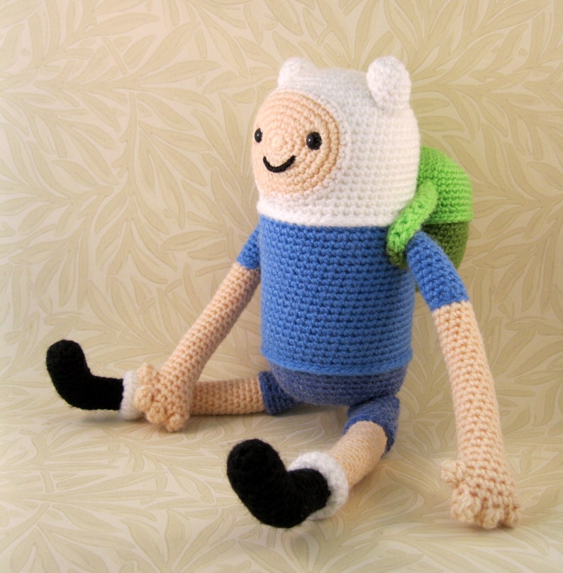 May include: A crocheted plush toy of Finn the Human from the animated series Adventure Time. Finn is wearing a blue shirt, white hat, and black shoes. He has a green backpack.