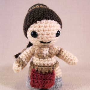 May include: Crocheted doll with brown hair, a brown and beige outfit, and a red skirt. The doll has black eyes and a smiling expression.