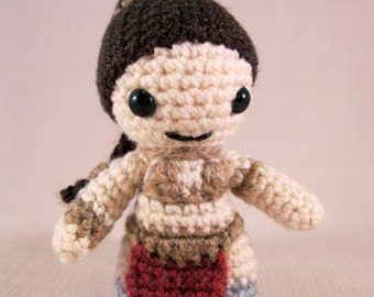 May include: Crocheted doll with brown hair, a brown and beige outfit, and a red skirt. The doll has black eyes and a smiling expression.