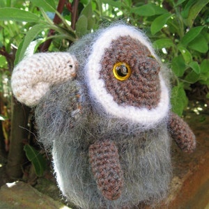 May include: A gray and brown crocheted stuffed animal with a white fur trim. The animal has large brown eyes with yellow pupils and a small white crocheted nose.