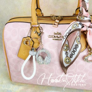 May include: A pink Coach handbag with a light brown trim and gold hardware. A white braided keychain with a rhinestone headphone charm hangs from the handle. A silk scarf with a chain and clock design is tied to the handle. The bag has a repeating pattern and the Coach logo.
