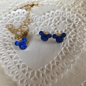 May include: A gold-colored necklace with a blue, glittery Mickey Mouse pendant and matching blue glitter stud earrings. The jewelry is displayed on a white, lace-like surface.