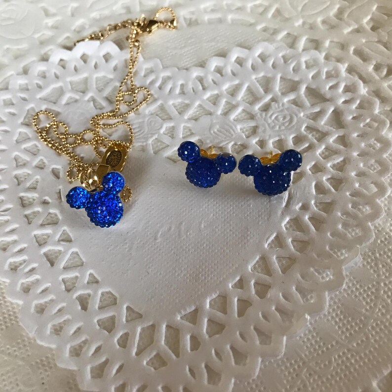 May include: A gold-colored necklace with a blue, glittery Mickey Mouse pendant and matching blue glitter stud earrings. The jewelry is displayed on a white, lace-like surface.