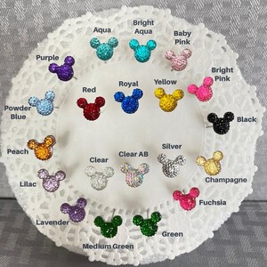 Puede incluir: Una colecci&oacute;n de pendientes de bot&oacute;n con forma de rat&oacute;n, incrustados con pedrer&iacute;a, en varios colores, incluyendo morado, aguamarina, rojo, amarillo y negro. Los pendientes se muestran sobre una superficie blanca y de encaje, con nombres de colores.