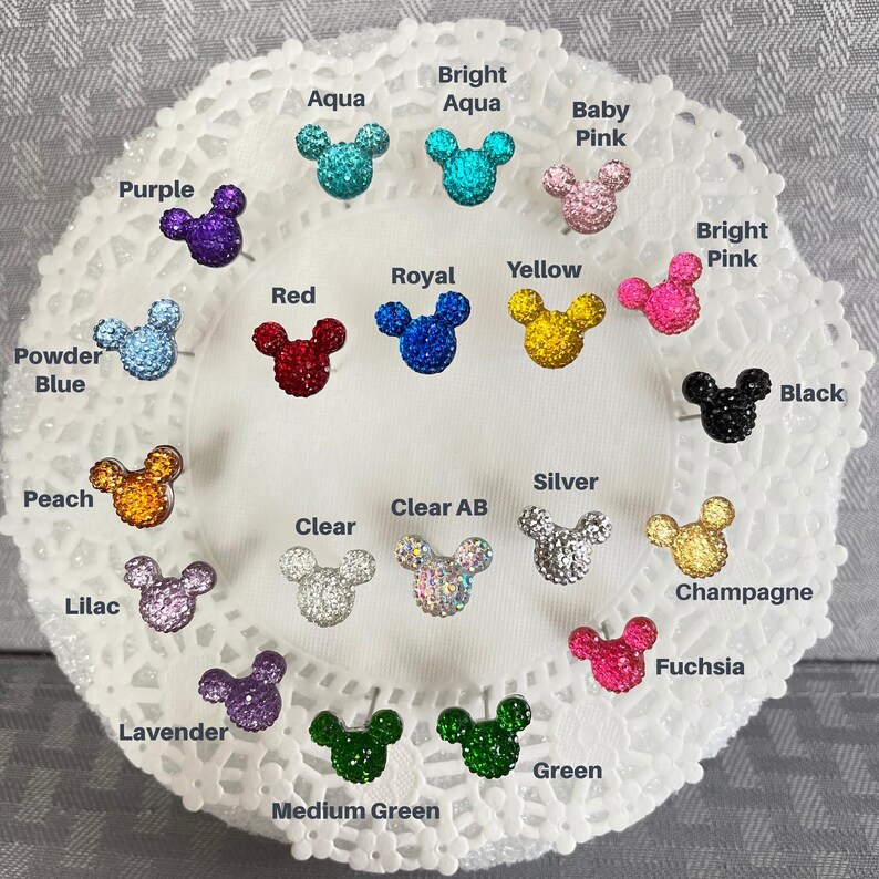 Puede incluir: Una colecci&oacute;n de pendientes de bot&oacute;n con forma de rat&oacute;n, incrustados con pedrer&iacute;a, en varios colores, incluyendo morado, aguamarina, rojo, amarillo y negro. Los pendientes se muestran sobre una superficie blanca y de encaje, con nombres de colores.