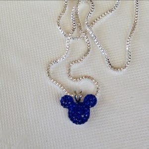 May include: A silver necklace with a pendant shaped like Mickey Mouse's head. The pendant is covered in small, sparkling blue crystals. The necklace is displayed on a white surface.