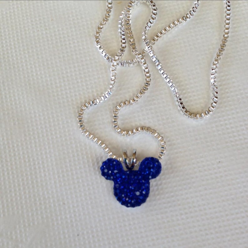 May include: A silver necklace with a pendant shaped like Mickey Mouse's head. The pendant is covered in small, sparkling blue crystals. The necklace is displayed on a white surface.