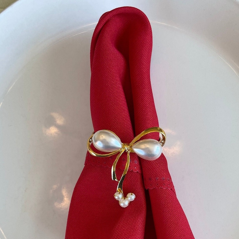 K&ouml;nnte beinhalten: Eine rote Serviette wird von einem goldfarbenen, schleifenf&ouml;rmigen Serviettenring gehalten. Der Serviettenring hat zwei gro&szlig;e, perlmuttfarbene Kunstperlen und drei kleinere Perlen. Die Serviette ist gefaltet und auf einem wei&szlig;en Teller platziert.