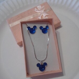 May include: A blue Mickey Mouse-shaped jewelry set, including a necklace and earrings, presented in a pink gift box with a ribbon. The jewelry has a sparkly, textured appearance, and the necklace features a silver chain.
