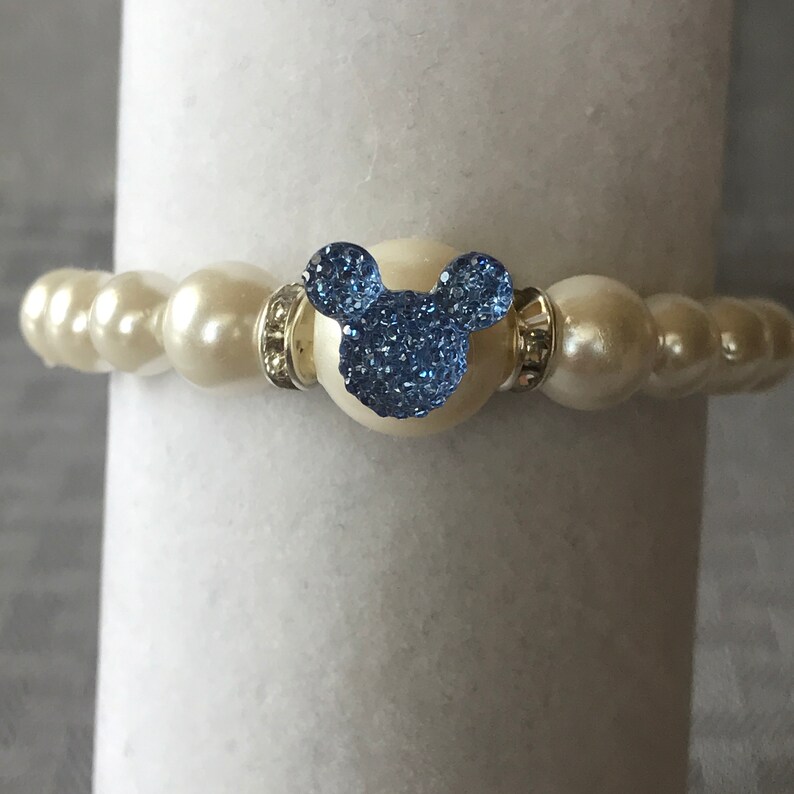 Puede incluir: Una pulsera de cuentas color crema con un dije azul en forma de Mickey Mouse. El dije est&aacute; incrustado con cristales azules brillantes. La pulsera est&aacute; hecha de perlas redondas grandes y detalles plateados.