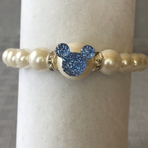 Puede incluir: Pulsera de cuentas color crema con un dije en forma de Mickey Mouse azul claro. El dije est&aacute; incrustado con cristales azules brillantes. La pulsera tiene detalles plateados.