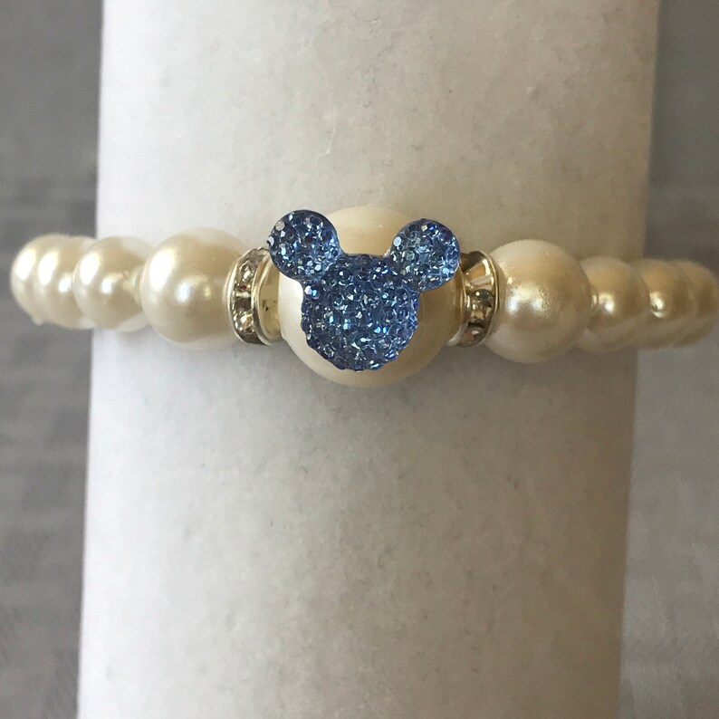 Puede incluir: Pulsera de cuentas color crema con un dije en forma de Mickey Mouse azul claro. El dije est&aacute; incrustado con cristales azules brillantes. La pulsera tiene detalles plateados.