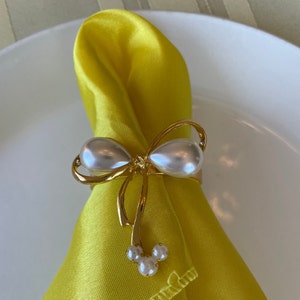 K&ouml;nnte beinhalten: Ein goldfarbener Serviettenring mit Schleifendesign, mit zwei gro&szlig;en, perlmuttartigen Kunstperlen. Der Ring h&auml;lt eine gefaltete gelbe Serviette, und eine Ansammlung von drei kleineren Perlen h&auml;ngt unterhalb der Schleife. Die Serviette liegt auf einem wei&szlig;en Teller.