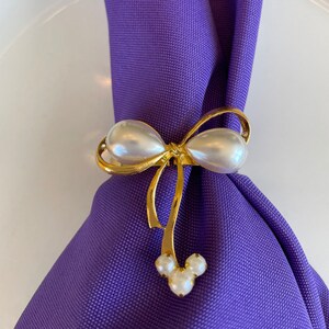 K&ouml;nnte beinhalten: Ein goldfarbener Serviettenring mit Schleifendesign, der gro&szlig;e, perlmuttartige Kunstperlen aufweist. Die Schleife h&auml;lt eine lila Serviette. Der Boden des Rings hat ein Mickey-Mouse-f&ouml;rmiges Design aus drei kleineren Perlen.