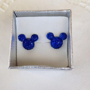 May include: A pair of blue crystal stud earrings in the shape of a mouse head, presented in a silver gift box with a white interior. The earrings are a vibrant blue and sparkle with many small crystals.
