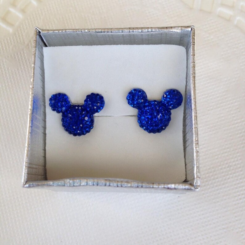 May include: A pair of blue crystal stud earrings in the shape of a mouse head, presented in a silver gift box with a white interior. The earrings are a vibrant blue and sparkle with many small crystals.