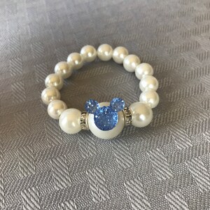 Puede incluir: Una pulsera de cuentas blancas con una gran cuenta central blanca con un dise&ntilde;o de Mickey Mouse azul brillante. La pulsera est&aacute; hecha de perlas falsas grandes y redondas y est&aacute; sobre un fondo gris.