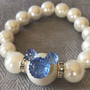Puede incluir: Una pulsera de perlas blancas con un dije azul en forma de Mickey Mouse. El dije est&aacute; hecho de peque&ntilde;os cristales azules brillantes. La pulsera est&aacute; ensartada en una banda el&aacute;stica.