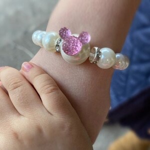 Puede incluir: Una pulsera de perlas blancas con un dije de Mickey Mouse rosa brillante. La pulsera presenta perlas grandes y redondas y detalles plateados. El dije es de color rosa brillante.