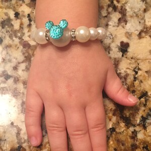 Puede incluir: Una pulsera de perlas blancas con un dije turquesa en forma de Mickey Mouse. El dije est&aacute; adornado con peque&ntilde;as piedras brillantes. La pulsera est&aacute; en la mano de un ni&ntilde;o.
