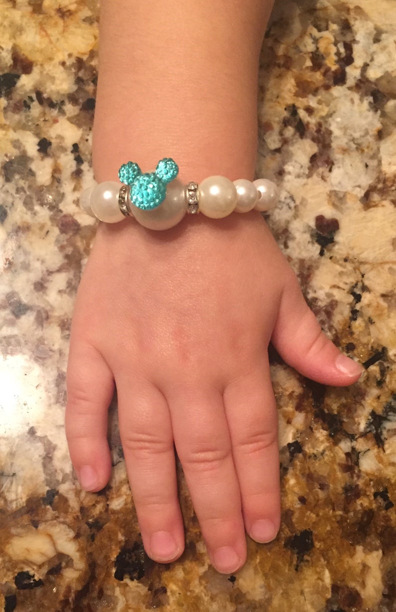 Puede incluir: Una pulsera de perlas blancas con un dije turquesa en forma de Mickey Mouse. El dije est&aacute; adornado con peque&ntilde;as piedras brillantes. La pulsera est&aacute; en la mano de un ni&ntilde;o.