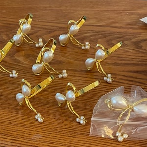 K&ouml;nnte beinhalten: Goldfarbene Metall-Serviettenringe mit Schleifendesign, jeweils mit Kunstperlen verziert. Die Schleife hat eine gro&szlig;e Perlenmitte und kleinere Perlencluster. Mehrere Ringe sind sichtbar, einige in einer durchsichtigen Plastikt&uuml;te.
